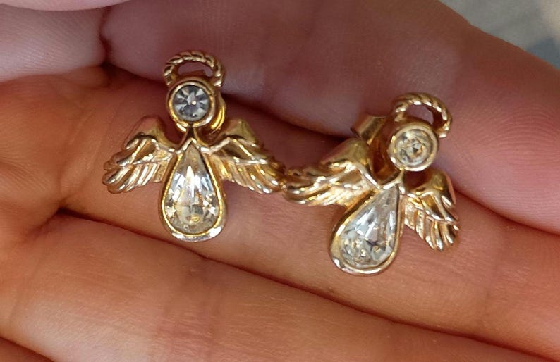 May include: A pair of gold-toned angel earrings. Each earring features a clear, teardrop-shaped crystal pendant and a smaller, round crystal at the head. The angels have detailed wings and halo accents.