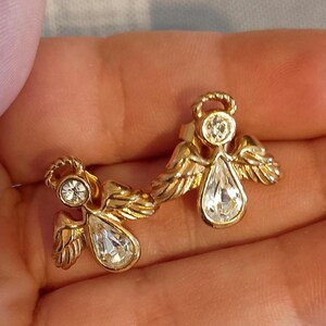 May include: A pair of gold-toned angel earrings. Each earring features an angel design with a halo, wings, and a teardrop-shaped clear crystal. The earrings are set against a neutral background.