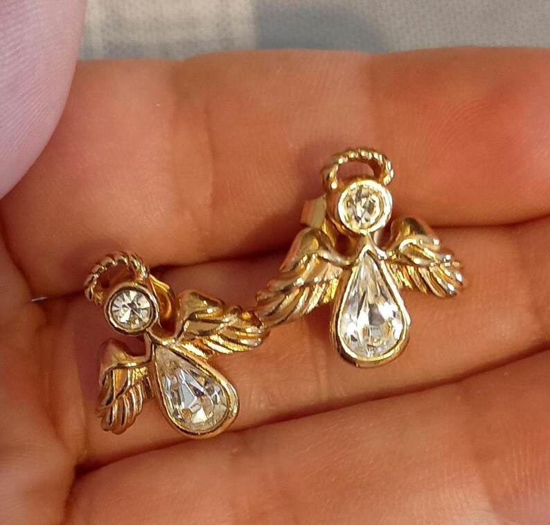 May include: A pair of gold-toned angel earrings. Each earring features an angel design with a halo, wings, and a teardrop-shaped clear crystal. The earrings are set against a neutral background.