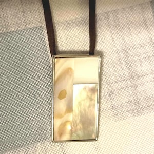 May include: A rectangular pendant necklace with a silver-colored frame. The pendant features a mosaic of cream, beige, and iridescent mother-of-pearl pieces. The necklace has a brown suede cord.
