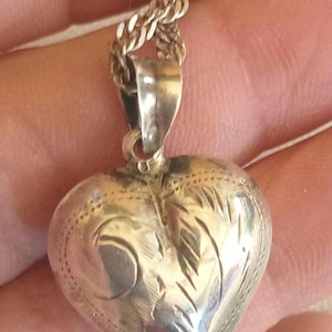 May include: Silver heart-shaped pendant necklace. The pendant has an engraved design and is attached to a chain. The heart is approximately 1 inch wide and 1 inch tall.