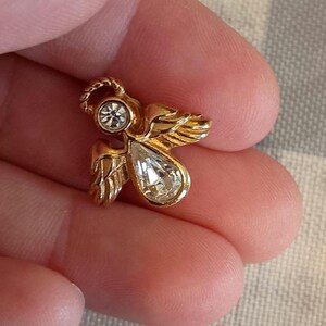 May include: A small, gold-colored angel pin with a clear, teardrop-shaped crystal and a smaller, round crystal. The angel has detailed wings and a halo. The pin is held in a person's hand.