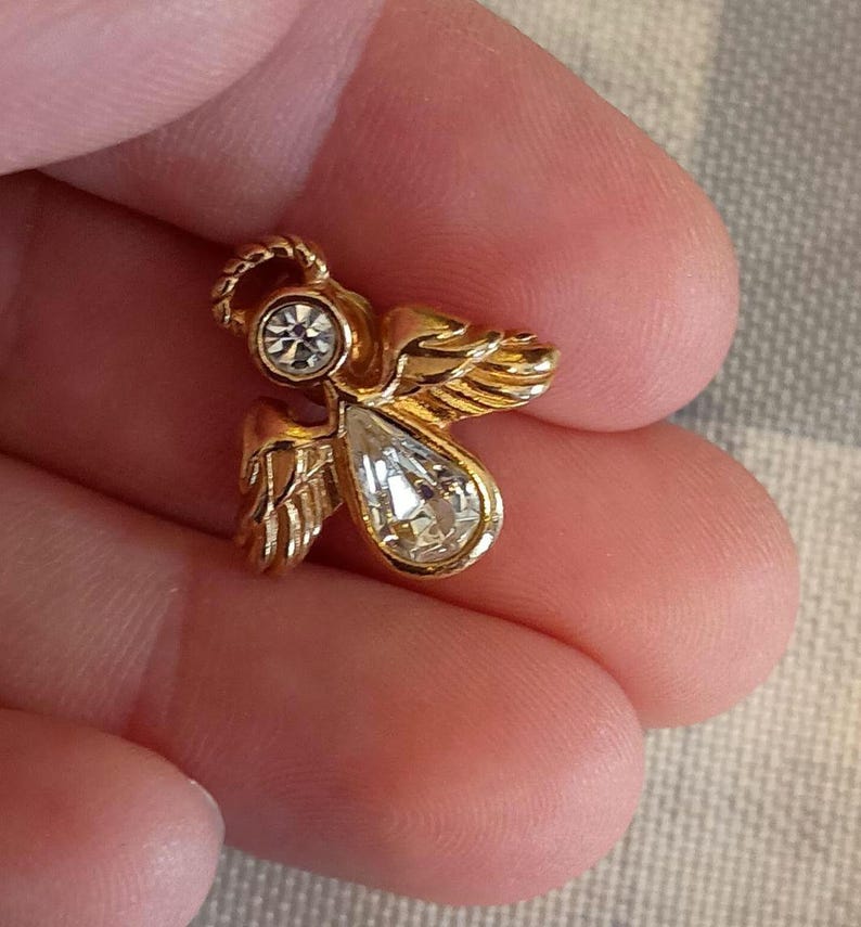 May include: A small, gold-colored angel pin with a clear, teardrop-shaped crystal and a smaller, round crystal. The angel has detailed wings and a halo. The pin is held in a person's hand.
