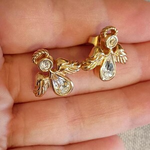 May include: A pair of gold-toned angel earrings. Each earring features an angel design with a halo, wings, and a teardrop-shaped crystal. The earrings are set against a neutral background.