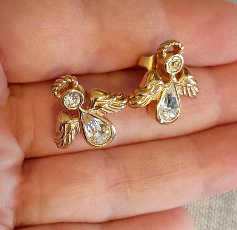 May include: A pair of gold-toned angel earrings. Each earring features an angel design with a halo, wings, and a teardrop-shaped crystal. The earrings are set against a neutral background.