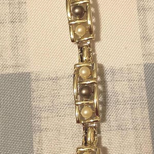 May include: Gold-toned bracelet featuring alternating cream and dark gray faux pearls. The bracelet has a rectangular link design with a clasp closure. A classic piece of jewelry.