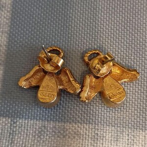 May include: A pair of gold-toned angel earrings. Each earring features an angel design with wings and a halo. The earrings have a post-back closure and are set against a textured background.
