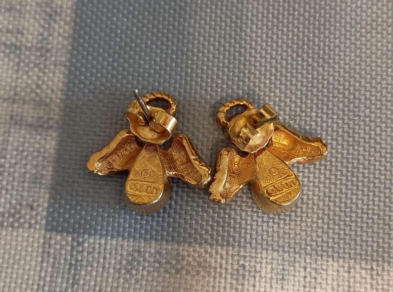 May include: A pair of gold-toned angel earrings. Each earring features an angel design with wings and a halo. The earrings have a post-back closure and are set against a textured background.