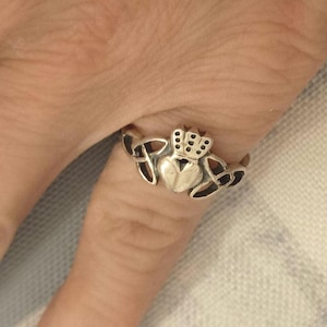 May include: A silver-toned Claddagh ring featuring a heart, crown, and hands design. The ring is worn on a finger, showcasing the intricate details of the jewelry. The heart is in the center, topped with a crown, and flanked by hands.