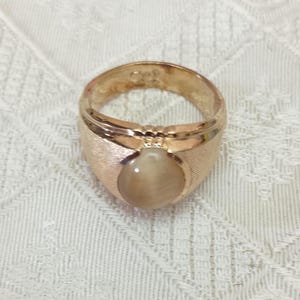 May include: A gold-toned ring featuring a central, smooth, round, light-brown cabochon gemstone. The band has a textured, brushed metal finish on the sides and a polished gold-toned setting around the stone. The interior of the band shows a hallmark.