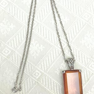 May include: Silver-toned necklace featuring a rectangular pendant with an amber-colored center. The pendant is framed with silver and has decorative detailing at the top. The chain is a delicate, linked design.