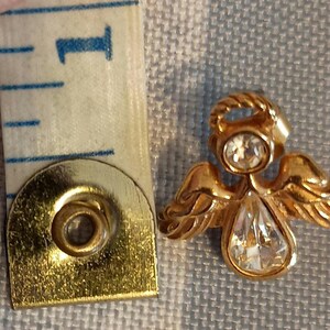 May include: A gold-colored angel pin with a clear, teardrop-shaped crystal pendant and a smaller, round crystal on the head. The angel's wings are detailed. A measuring tape is next to the pin, showing the 1-inch mark.