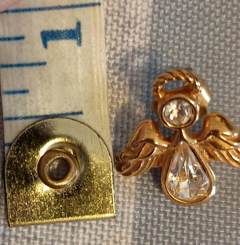 May include: A gold-colored angel pin with a clear, teardrop-shaped crystal pendant and a smaller, round crystal on the head. The angel's wings are detailed. A measuring tape is next to the pin, showing the 1-inch mark.