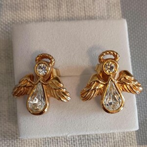 May include: A pair of gold-toned angel stud earrings. Each earring features a halo, a round clear stone, and a pear-shaped clear stone. The wings are detailed with feather-like textures. The earrings are displayed on a white surface.