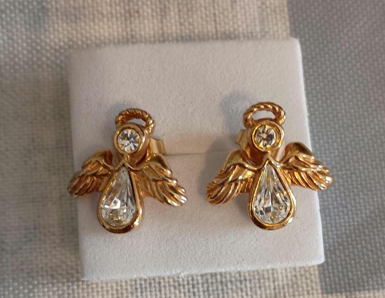 May include: A pair of gold-toned angel stud earrings. Each earring features a halo, a round clear stone, and a pear-shaped clear stone. The wings are detailed with feather-like textures. The earrings are displayed on a white surface.