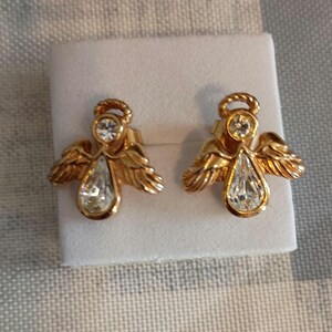 May include: A pair of gold-toned angel earrings. Each earring features an angel design with a clear, teardrop-shaped crystal pendant and a smaller round crystal at the top. The wings are detailed, and the earrings are presented on a white surface.