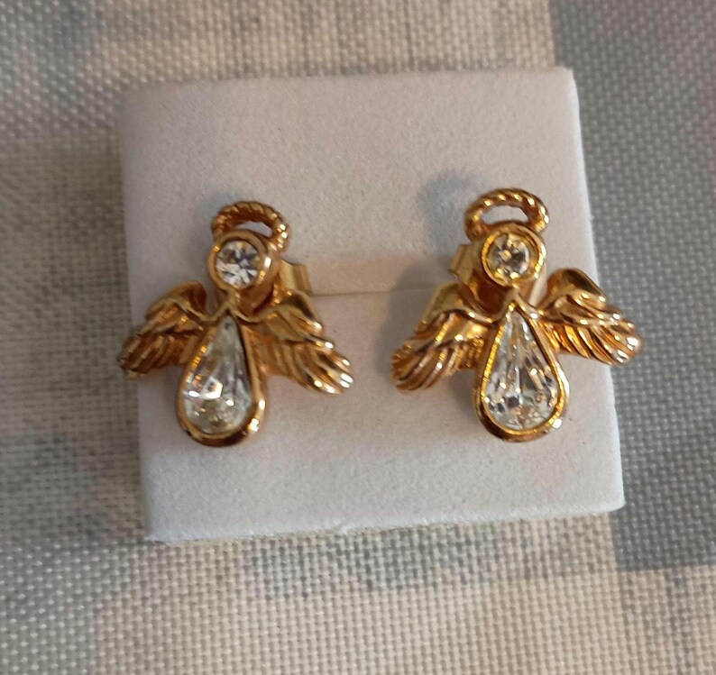 May include: A pair of gold-toned angel earrings. Each earring features an angel design with a clear, teardrop-shaped crystal pendant and a smaller round crystal at the top. The wings are detailed, and the earrings are presented on a white surface.