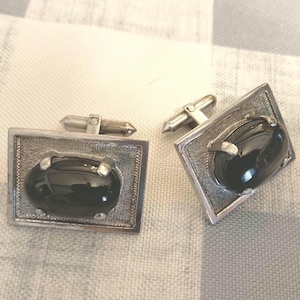 May include: A pair of silver cufflinks with a rectangular frame and an oval, black stone center. The cufflinks have a textured border and a silver post for fastening. The background is a light gray and white checkered pattern.