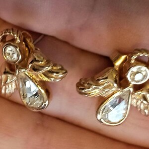 May include: A pair of gold-toned angel earrings with clear, teardrop-shaped crystals. The earrings feature detailed wings and halos, creating a vintage or celestial aesthetic. The earrings are shown against a neutral background.