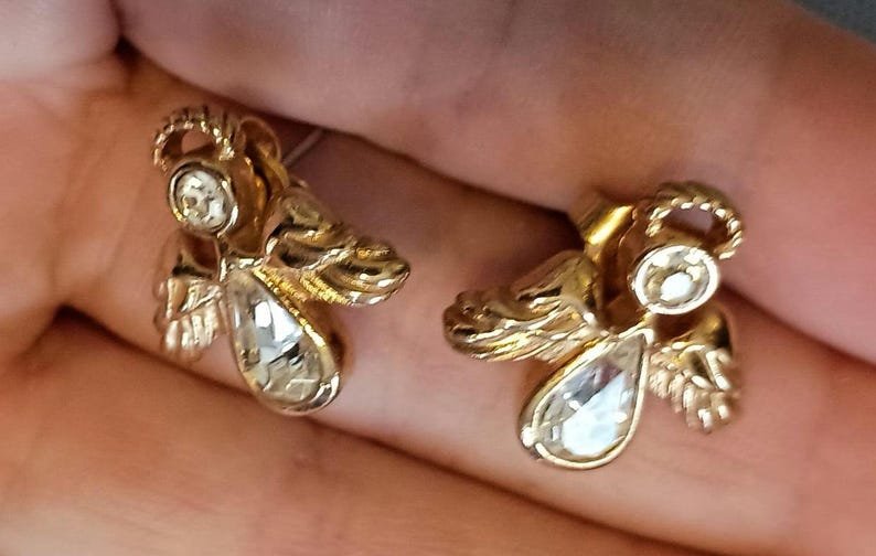 May include: A pair of gold-toned angel earrings with clear, teardrop-shaped crystals. The earrings feature detailed wings and halos, creating a vintage or celestial aesthetic. The earrings are shown against a neutral background.