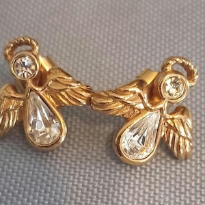 May include: A pair of gold-toned angel earrings. Each earring features a halo, a round clear stone, and a teardrop-shaped clear stone. The wings are detailed with feather-like textures.