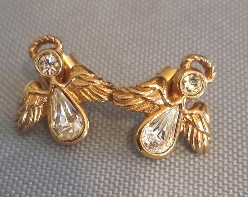 May include: A pair of gold-toned angel earrings. Each earring features a halo, a round clear stone, and a teardrop-shaped clear stone. The wings are detailed with feather-like textures.