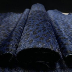 Könnte beinhalten: Ein Stück blau-schwarzes Rindsleder mit Tierfellmuster und einer kurzhaarigen, strukturierten Oberfläche. Das Material ist in der Mitte gefaltet und zeigt ein sich wiederholendes dunkles Fleckenmuster auf einem leuchtend blauen Grund.