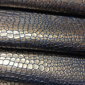 Op de afbeelding: Een stapel gevouwen stof met een metallic brons- en goudkleurige krokodillenleertextuur. Het reliëfpatroon bestaat uit onregelmatige, verhoogde schubben met diepblauwe kieren tussen elke vorm, wat zorgt voor een multidimensionale uitstraling.