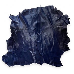 Pode incluir: Uma peça única e irregular de couro azul-marinho escuro com uma superfície lisa e ligeiramente reflexiva. O material apresenta bordas naturais e uma textura macia e flexível com dobras e vincos subtis em toda a sua superfície.