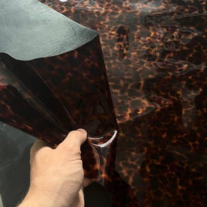 Può includere: Un foglio di materiale flessibile caratterizzato da un motivo tartarugato lucido, nei toni del marrone scuro e del nero. La superficie è liscia e riflettente, con un angolo ripiegato che rivela un retro nero opaco e il profilo sottile.