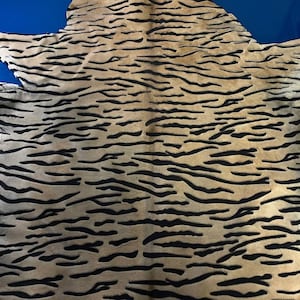 Puede incluir: Una alfombra de piel animal de forma irregular con una base de color beige y un patrón repetitivo de rayas negras horizontales y onduladas en toda la superficie. Los bordes de la piel son desiguales y dentados, siguiendo los contornos naturales.