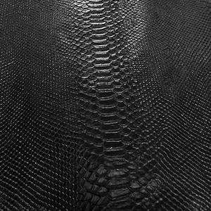 Peut inclure: Un matériau noir présentant un motif texturé et gaufré imitant l'apparence d'écailles de serpent. La surface affiche une disposition répétitive et complexe de petites écailles en relief en forme de losange avec une arête centrale.