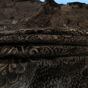 May include: A dark brown fabric featuring an intricate, raised floral pattern with a textured, perforated surface. The material is folded in layers, showcasing a deep, monochromatic color palette with varying light reflections across the embossed design.