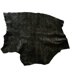 Pode incluir: Uma peça de couro preto com uma textura de superfície rachada. O material tem uma forma orgânica irregular com dobras visíveis no seu acabamento mate escuro, exibindo uma rede distinta de linhas finas e claras por toda a superfície.