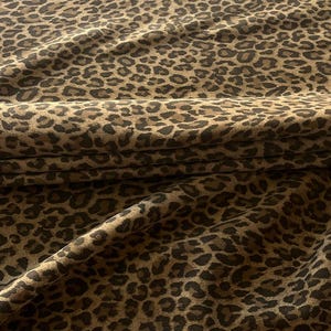 Puede incluir: Una pieza de tela doblada con un patrón de leopardo repetitivo. El diseño consiste en manchas irregulares de color marrón oscuro y negro sobre un fondo de color tostado y marrón claro, creando una apariencia texturizada con pliegues visibles.