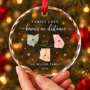 May include: A circular clear glass ornament with faceted edges, featuring the text "FAMILY LOVE knows no distance", "THE MILLER FAMILY", and "2026". It displays three state maps connected by dotted lines with red hearts and white snowflakes.