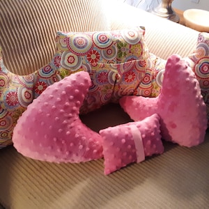 May include: Five pillows, two are pink and three are multi-colored with a circular pattern. The pink pillows are shaped like a crescent moon. The multi-colored pillows are rectangular.