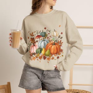 Könnte beinhalten: Ein beigefarbenes Sweatshirt mit Rundhalsausschnitt und einem zentralen Grafikdruck aus verschiedenen Kürbissen, Zierkürbissen, Pilzen und Herbstblättern in Orange-, Grün-, Blau- und Brauntönen. Das langärmelige Kleidungsstück hat einen gerippten Ausschnitt, Bündchen und Saum.