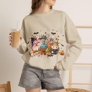 Könnte beinhalten: Ein beigefarbenes Sweatshirt mit Rundhalsausschnitt und einem bunten Grafikdruck aus verschiedenen lächelnden Kürbissen, Pilzen, Fledermäusen und einem Spinnennetz. Das Design enthält Herbstblätter, kleine Sterne und ein Totenkopfmotiv.