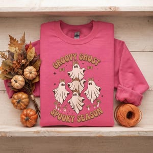 Könnte beinhalten: Ein leuchtend pinkfarbenes Sweatshirt mit Rundhalsausschnitt und einer Grafik von vier weißen Geistern mit braun gemusterten Schleifen, umgeben von kleinen Pilzen und Sternen, mit dem Text "GROOVY GHOST SPOOKY SEASON" in brauner Retro-Schrift.