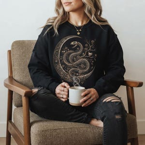 Peut inclure: Un sweat-shirt noir à manches longues arborant un grand motif détaillé représentant un squelette de serpent enroulé parmi des branches et des roses, le tout sur fond de croissant de lune. Le design est décliné en tons bronze et or.