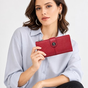 May include: A rectangular red wallet with a textured finish, featuring a silver-toned metal turn-lock closure on the front flap and a silver zip pull on the side. The wallet is held horizontally, showcasing its slim design and hardware details.
