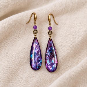 May include: A pair of drop earrings featuring teardrop-shaped pendants with a purple iridescent shell inlay. Each earring has a small round purple bead and a metallic rose-shaped accent attached to a gold-toned hook for pierced ears.