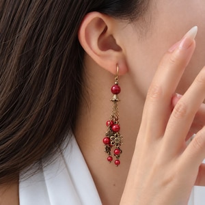 May include: A dangling earring featuring a single red bead at the top, connected to a cluster of bronze-toned chains. The chains are adorned with smaller red beads and a small, intricate bronze flower charm at the bottom of the design.