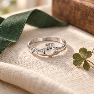 May include: A silver-toned Claddagh ring featuring a central heart shape topped with a small crown, flanked by two hands that meet at the centre. The band is smooth and polished, showcasing the traditional design elements of the iconic ring style.