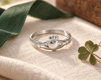 Può includere: Un anello Claddagh color argento caratterizzato da un cuore centrale sormontato da una piccola corona, affiancato da due mani che si incontrano al centro. La fascia è liscia e lucida, mettendo in risalto gli elementi di design tradizionali.