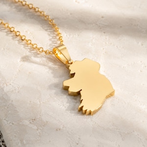 May include: A gold-toned necklace featuring a pendant shaped like the island of Ireland. The pendant is suspended from a delicate gold-toned chain link. The surface of the metal is smooth and reflective, catching the light to highlight the distinct geographical outline of the landmass.
