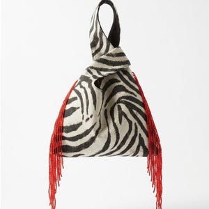May include: A triangular handbag featuring a black and white zebra stripe pattern with a single knotted handle. The two side edges of the bag are adorned with long, vibrant red beaded fringe that hangs down from the top to the bottom corners.