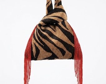 May include: A knot-handle handbag featuring a brown and black tiger stripe pattern on a textured fabric. The bag is accented with long, vibrant red beaded fringe hanging from both sides, creating a distinct contrast against the animal print.