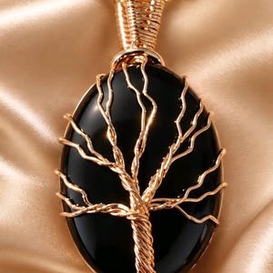 May include: A pendant necklace featuring a smooth, oval-shaped black stone wrapped in gold-coloured wire. The wire is intricately twisted to form the shape of a tree with branches extending across the surface of the stone, suspended from a gold chain.